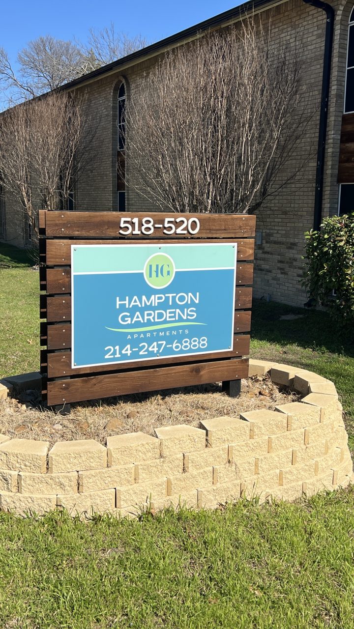 a sign for an apartment in front of a grassy area at The Hampton Gardens